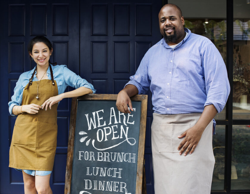 Cheerful woman and man business owners leaning on an a-frame blackboard sign.