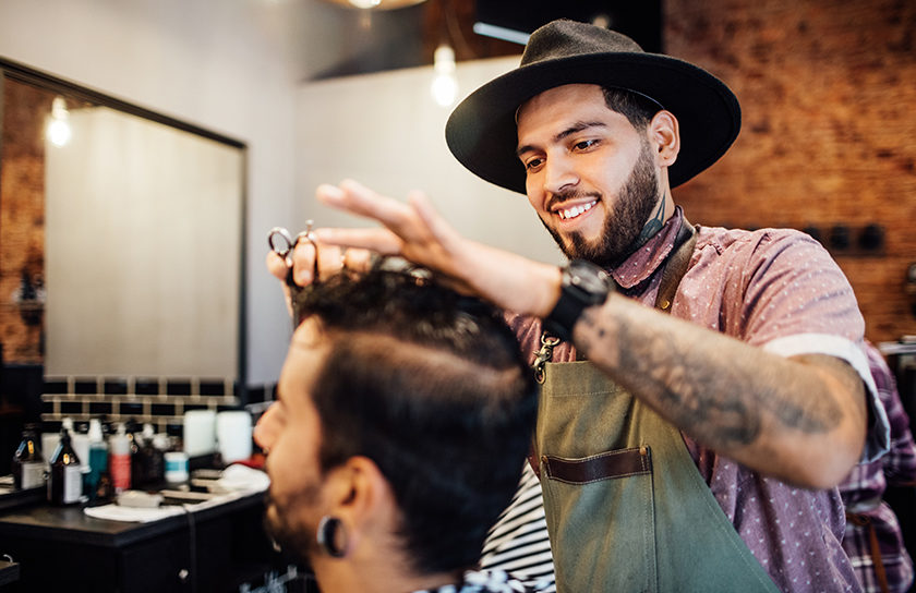 smiling man in hat cutting another mans hair