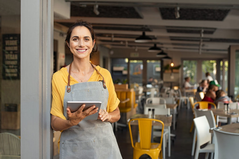 Happy employee standing at doorway of her store holding digital tablet.